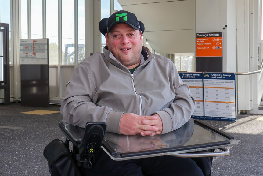 a man in a wheelchair smiling at the camera at a train station wearing a black cap and a grey hoodie