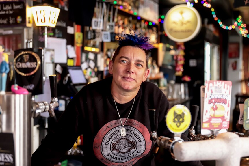 Woman with purple hair and black t-shirt stands behind pub bar with beer taps in foreground