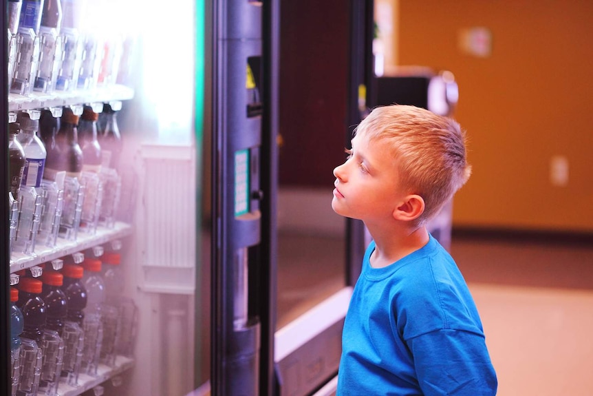 A a small blonde child looks longingly at a vending machine.