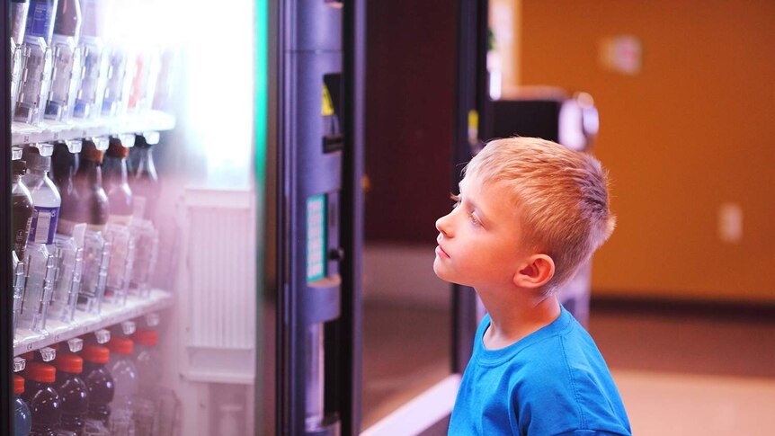A a small blonde child looks longingly at a vending machine.