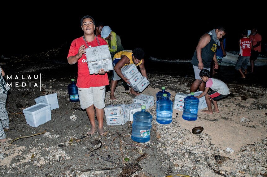 Young boy in red shirt holds water supplies on beach, surrounded by people collecting more supplies after tsunami. 
