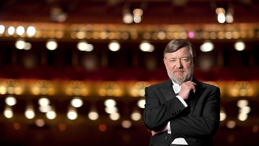 A man in a black formal jacket  and white shirt in front of a dark theatre