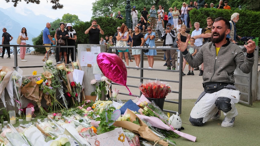 A young man of Middle-Eastern descent cries as he kneels in front of floral tributes left at a playground, while people watch.