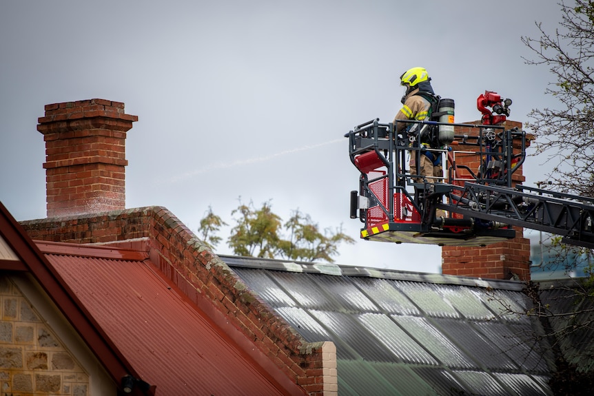 A firefighter standing on a platform of a crane directs a hose of water at a roof of a house