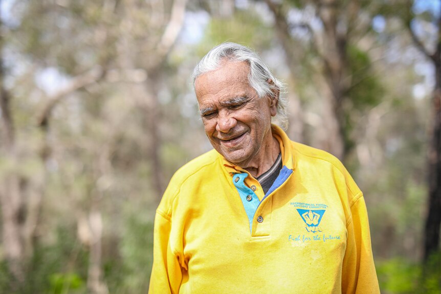 An Aboriginal man in a yellow shirt smiles and looks towards the ground, blurred trees in the background.