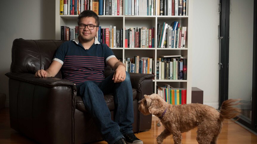A 31-year-old Asian man sits on a brown leather armchair in front of a book case while brown poodle plays in foreground
