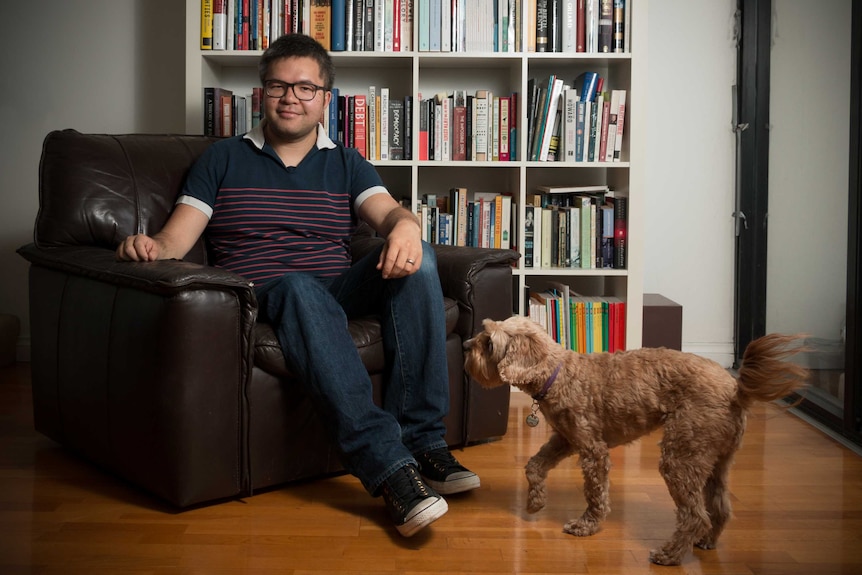 A 31-year-old Asian man sits on a brown leather armchair in front of a book case while brown poodle plays in foreground