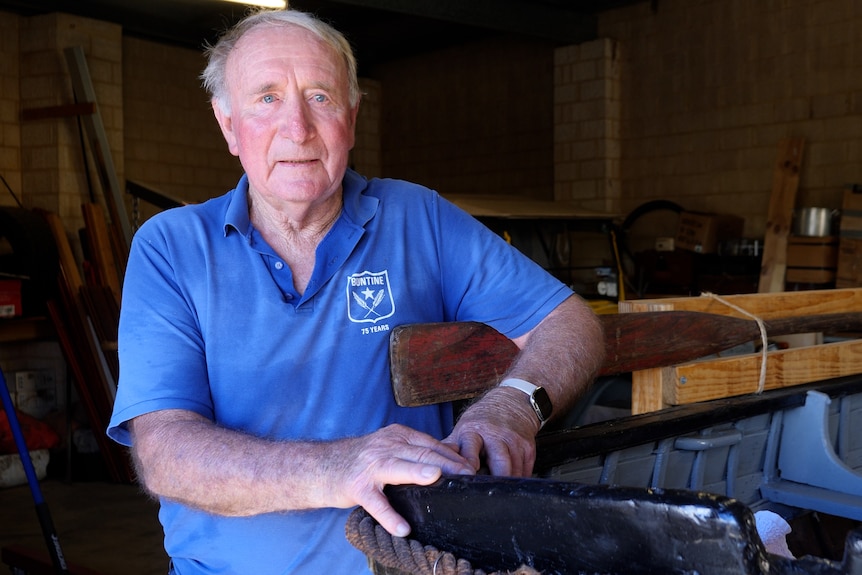 An elderly man in a shed with his hands resting on the corner of an old boat.