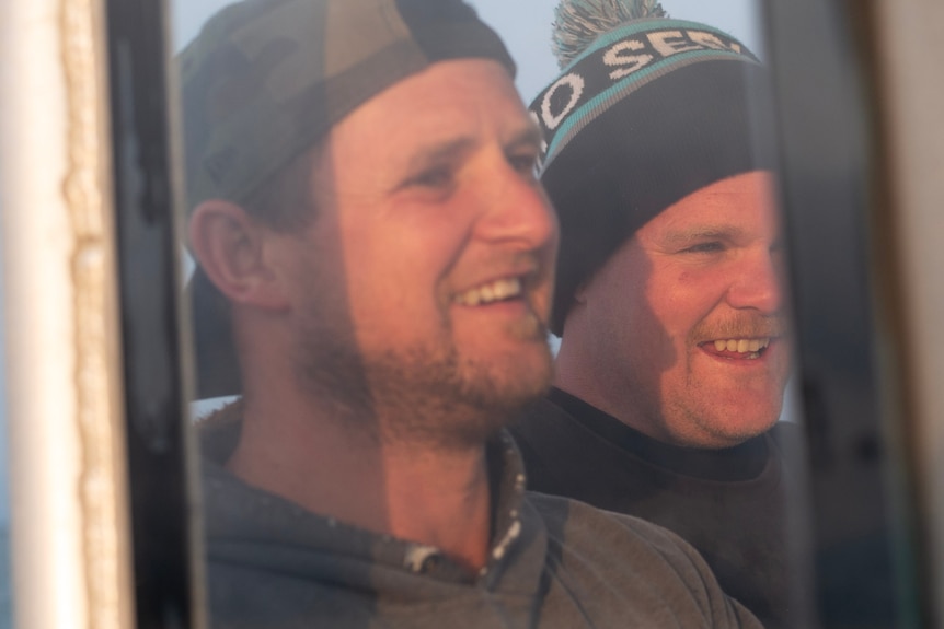 two men's faces fill the frame as they are seen through a salt-covered window on a boat.