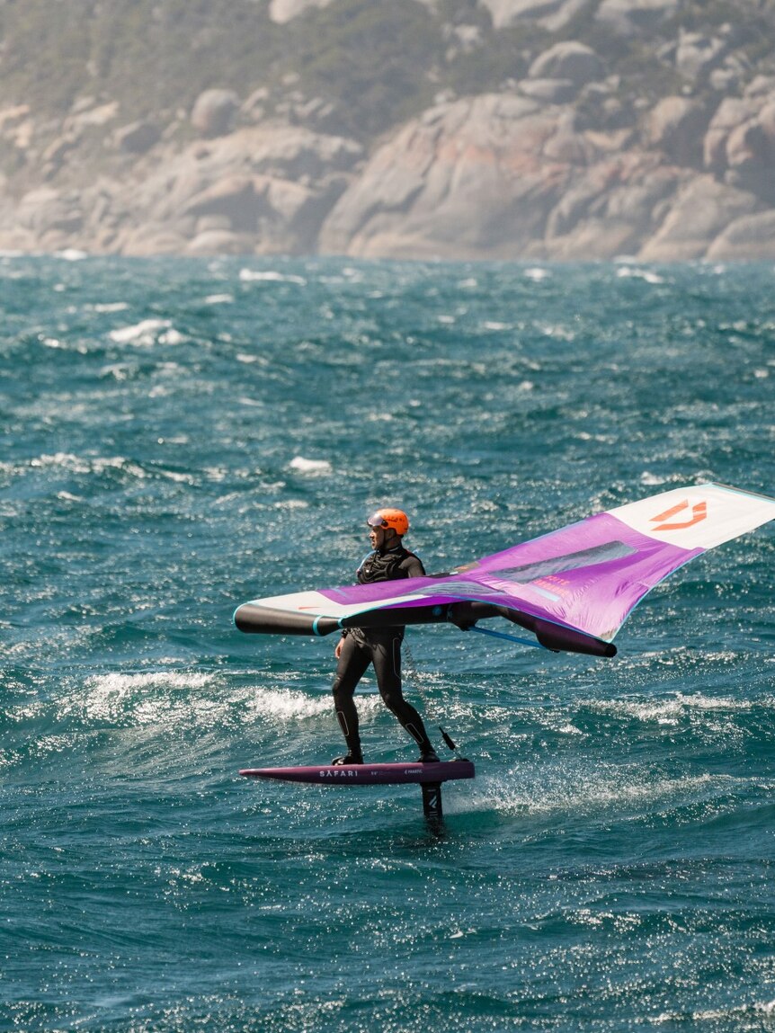 A man seen on a windsurfer in the ocean, with rocks in the background