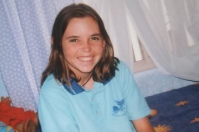 A smiling Hayley Dodd sits on a bed wearing a light blue school polo shirt.