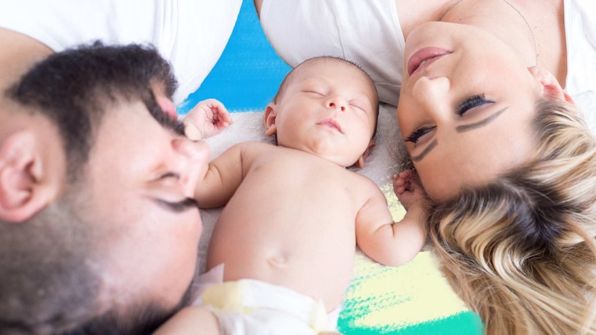 A mum and dad lie on a bed with their newborn baby sleeping between them for a story about postnatal depletion.