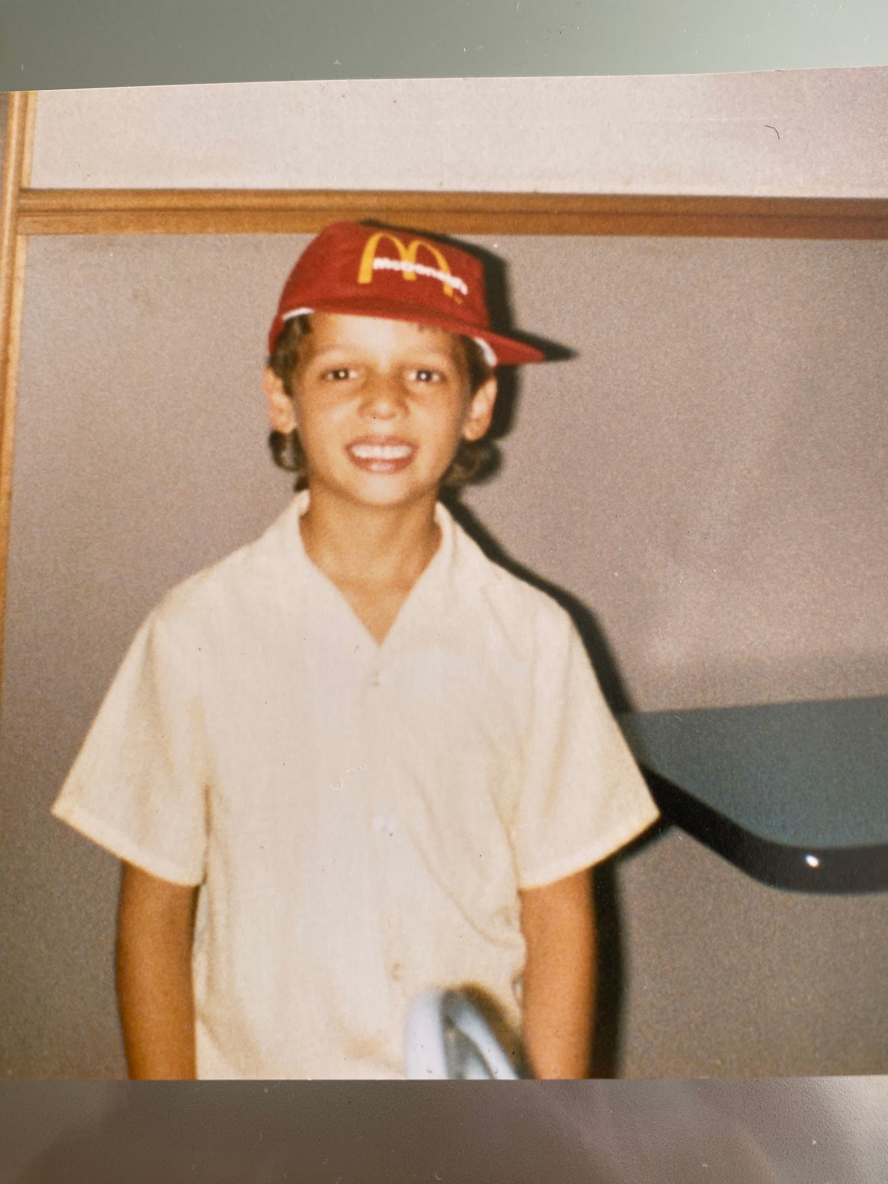 A printed photo of young Maltese Australian boy smiling and wearing a pale yellow shirt and a red McDonalds cap.