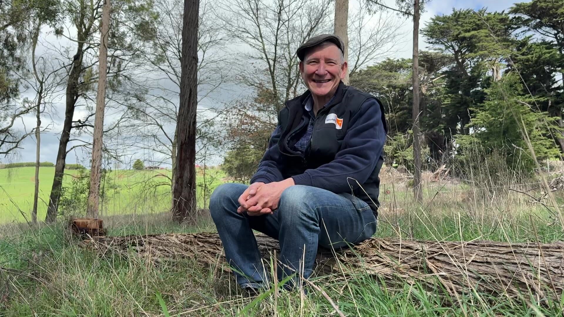 A man sits on a log in a paddock, surrounded by trees and long grasses. 