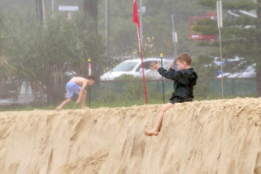 A child in a wind breaker throws sand in the air as he sits on the edge of scarping at Burleigh in rainy weather.