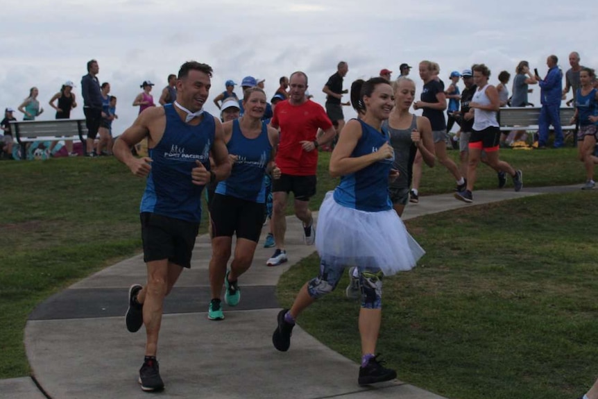 A man and woman in running gear with white, wedding accessories run on a path park ahead of a group of other runners.