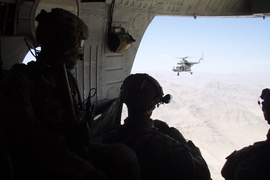 Soldiers in a helicopter mid-flight look out to another helicopter.