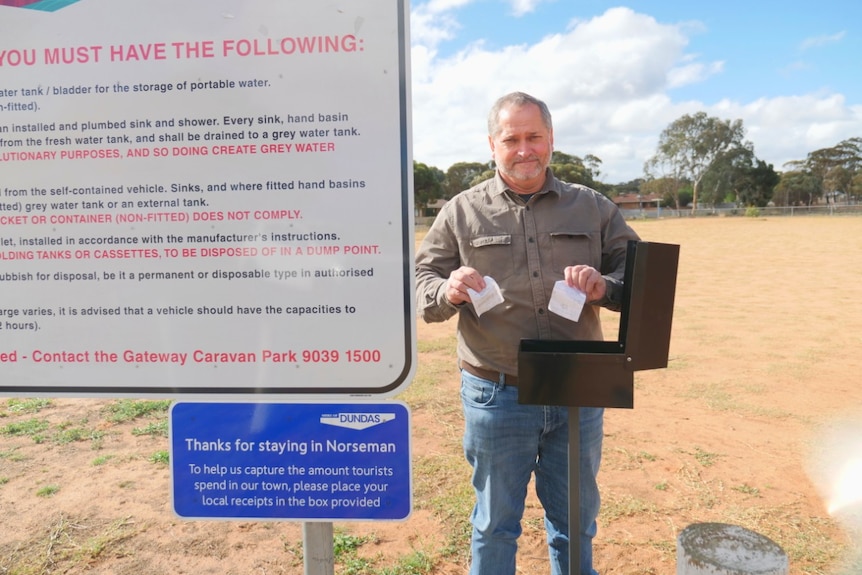 A man wearing jeans and a shirt stands in front of a feedback box at a caravan park holding two small white squares of paper.