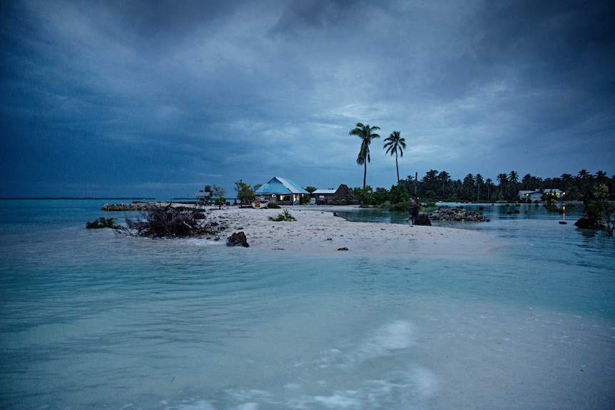 water comes very close to either side of a small sand bank. A blue house and palm trees are located in the distance