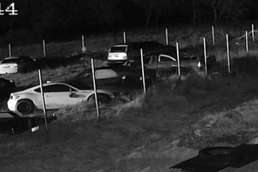 CCTV footage of a car yard, in the distance a hooded figure moves past cars