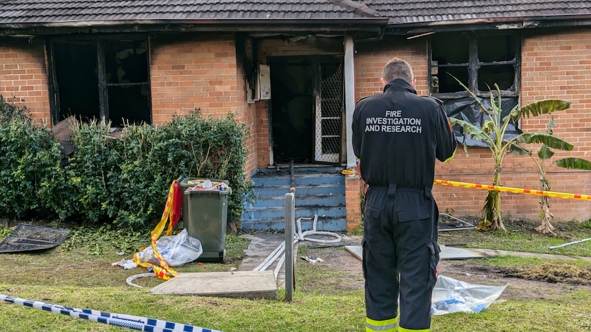 A man in a police investigations jumpsuit stands with his back to the camera in front of a burnt out home.