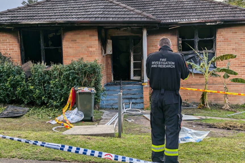 A man in a police investigations jumpsuit stands with his back to the camera in front of a burnt out home.