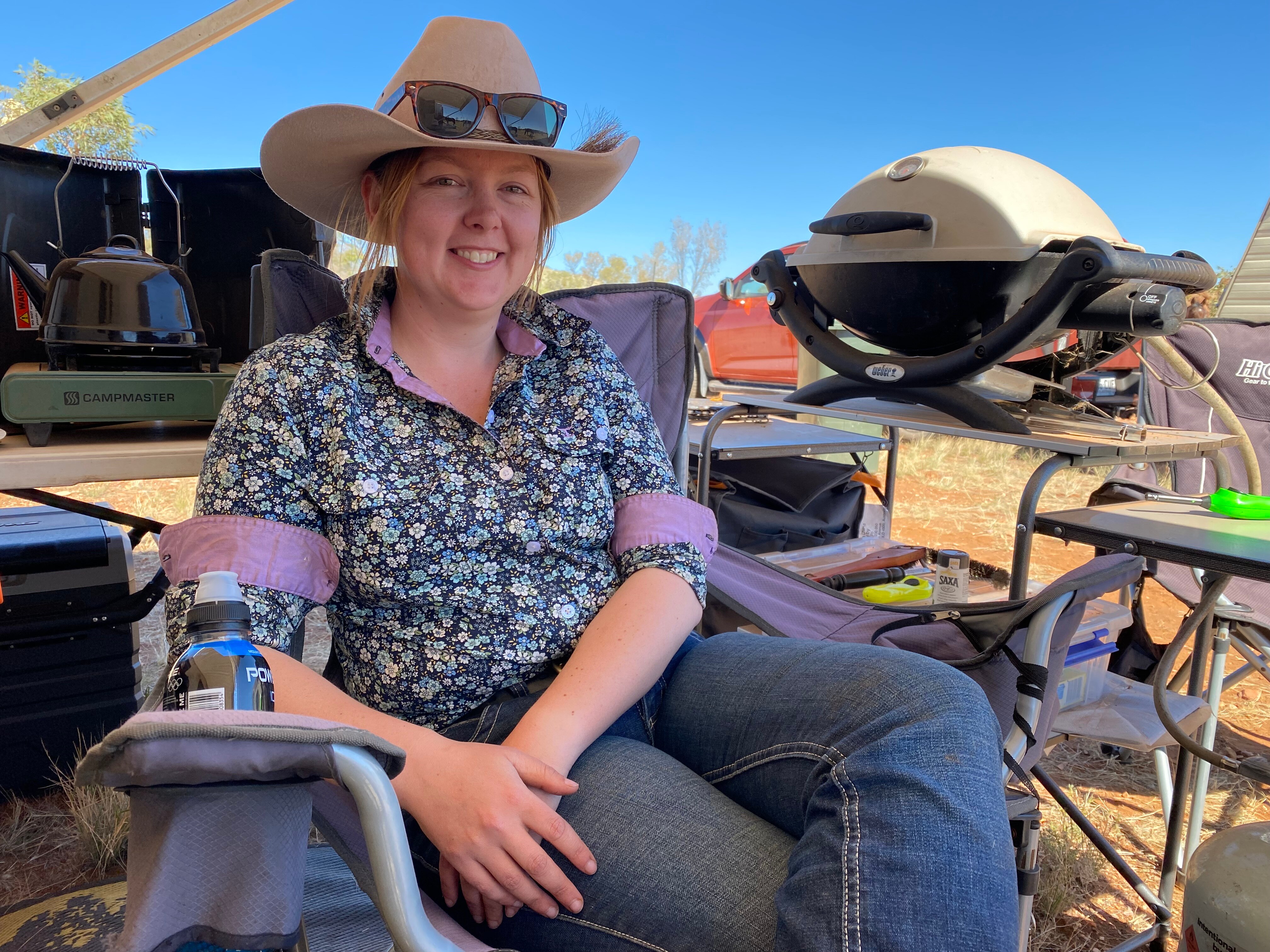 A young woman wearing a floral collared shirt, cowgirl hat and sunglasses rests in a campchair and smiles at the camera.
