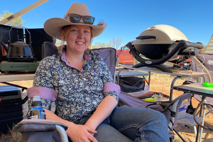 A young woman wearing a floral collared shirt, cowgirl hat and sunglasses rests in a campchair and smiles at the camera.