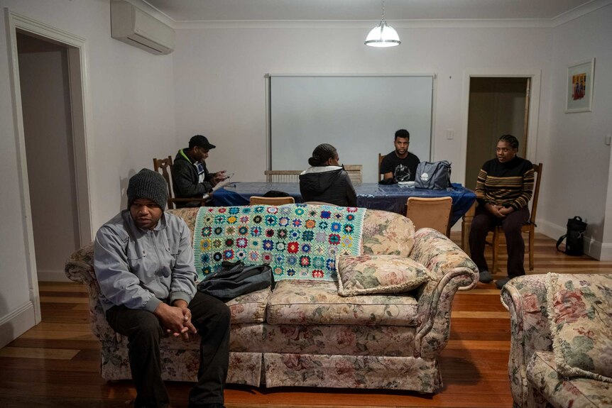 Four people sit at a dining table in the background, while a man in a beanie sits on a couch in the foreground.