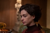 White woman with short brunette hair wears a red long-sleeved shirt and sits looking despondent at a dinner table.