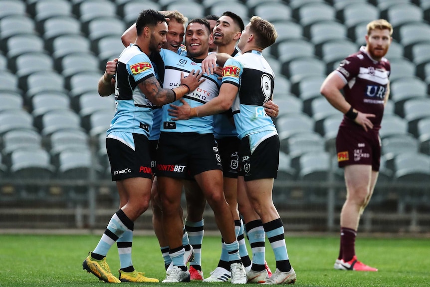 Cronulla NRL players surround a teammate to celebrate his try as a Manly opponent looks on in the background.