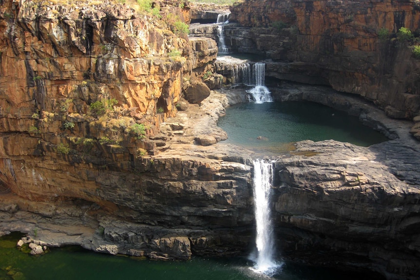 A wide shot of the Mitchell Falls in WA's Kimberley, showing a series of waterfalls on a steep, tiered cliff face with a pond.
