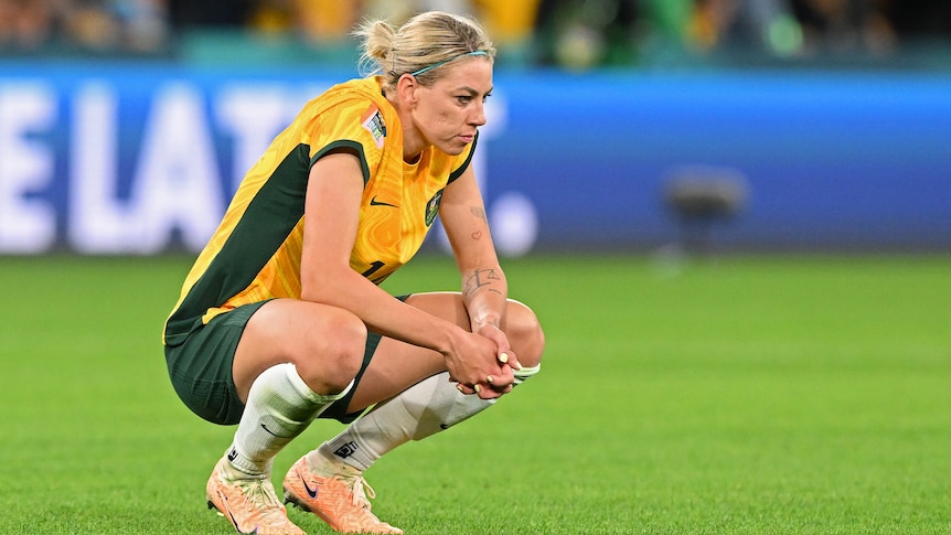A soccer player wearing yellow and green sits on her haunches after a game