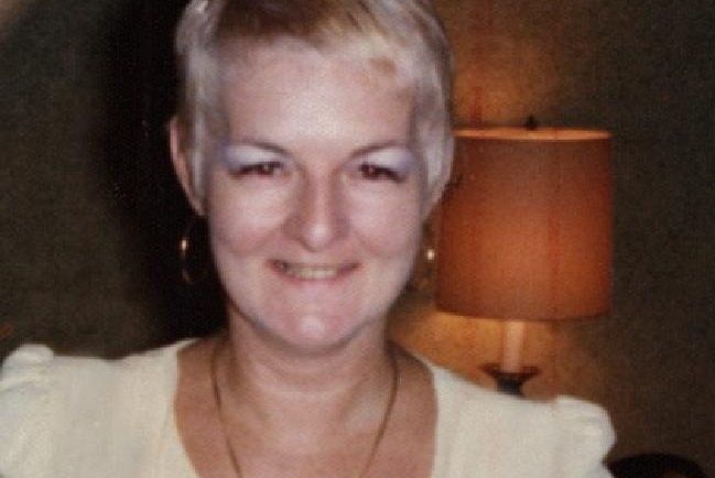 Shirley Finn stands with her hands on her hips in a white dress smiling for the camera.