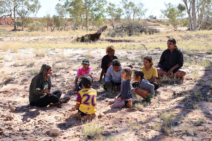Young woman surrounded by children, all sitting in a clearing in the bush.  