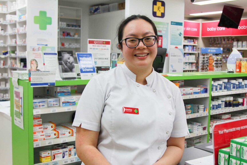 A smiling pharmacist stands behind a counter. There are shelves of drugs behind her