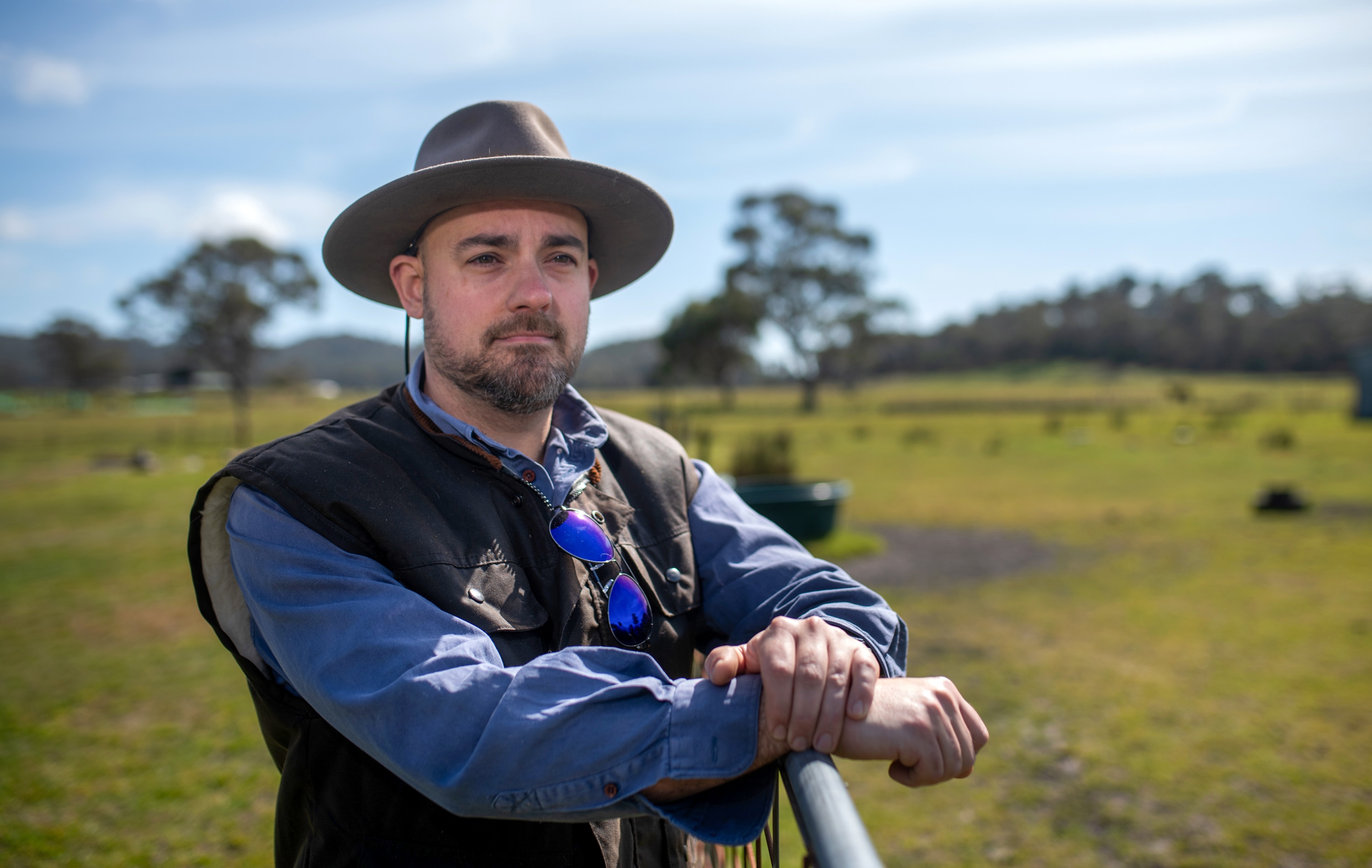 Ryan leans on a metal fence wearing a hat and looking over his property with distant trees.