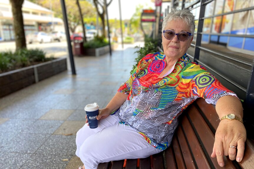 A woman with very colourful shirt holds a coffee cup while she sits on a public bench.