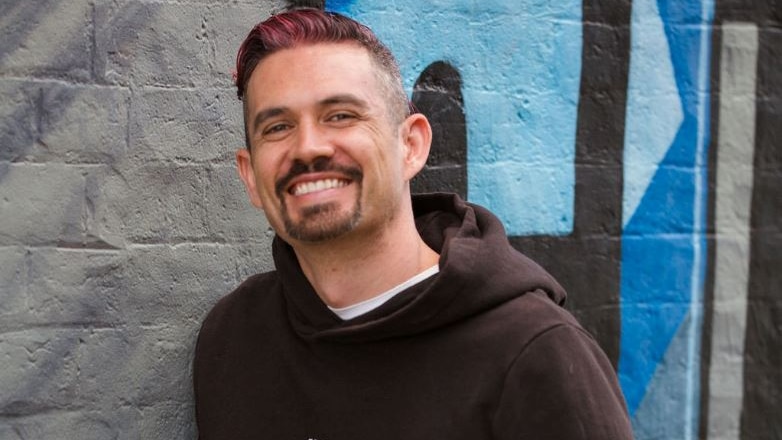 light skinned man with dark coloured short cut hair, smiling and wearing a black hoody. standing in front of a graffit wall  