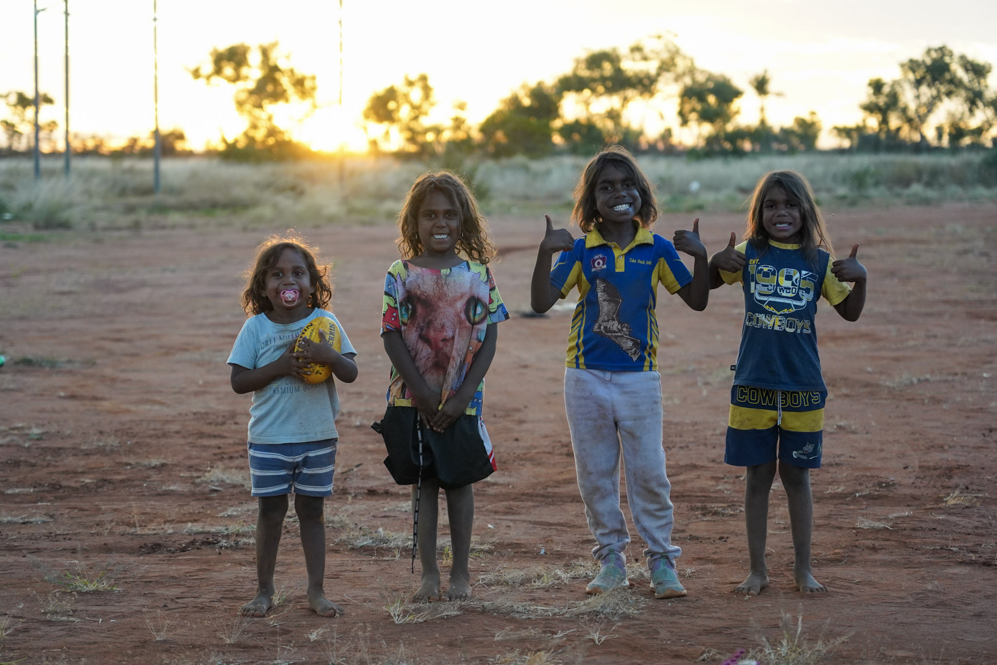 Four young Indigenous children standing on red dirt oval