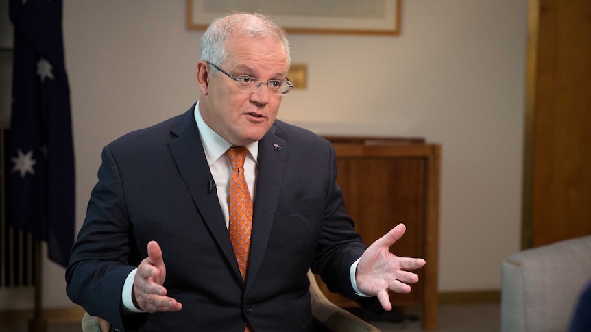 Mr Morrison is sitting in a chair and gesturing with his hands, looking right of frame. He's wearing an orange tie.