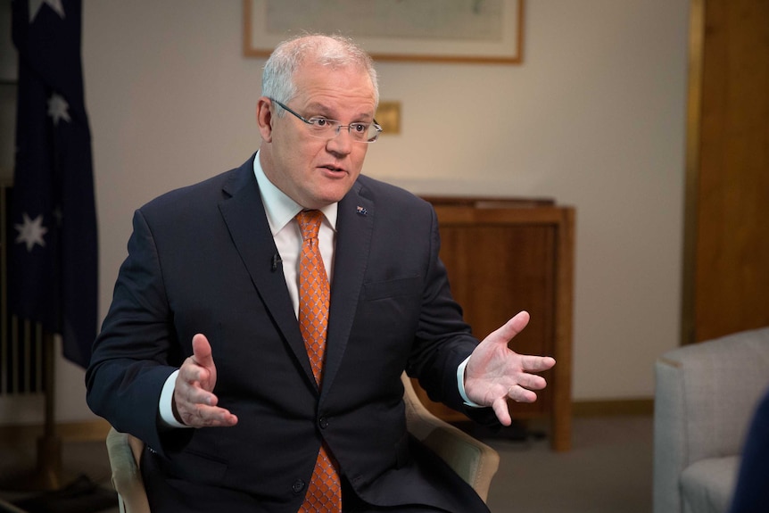 Mr Morrison is sitting in a chair and gesturing with his hands, looking right of frame. He's wearing an orange tie.