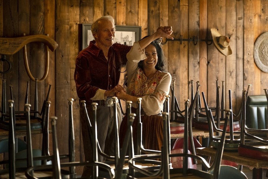 A 90-something man and a 50-something woman smile and widely as they dance together in an empty restaurant space