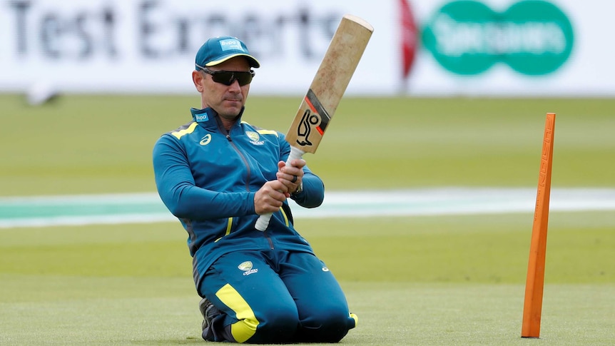 Australia cricket coach Justin Langer, in training gear, kneels in front of a practice stump while holding a Kookaburra bat.