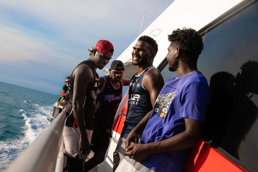 Four men stand on the side of a boat as it cruises along the water