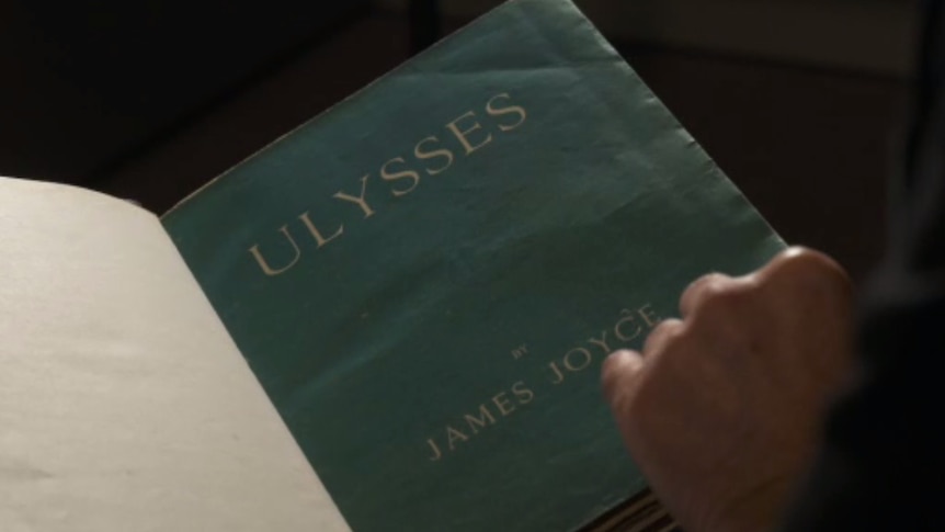 A hand holds an old blue book. The cover says Ulysses by James Joyce.