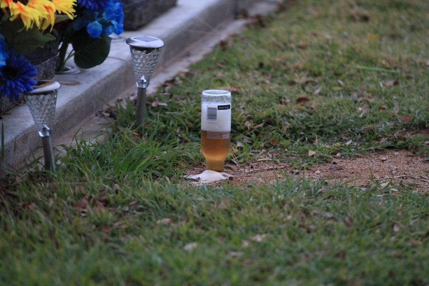 A bottle of beer stuck into the earth upside down next to a grave stone.