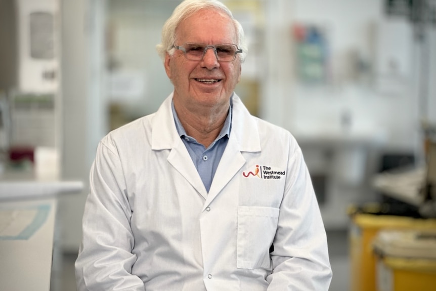 A man with white hair and a white lab coat smiles in a lab room
