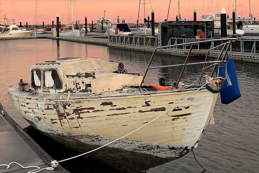 A dilapidated yacht rests in centimetres of water at a boat ramp with a sunrise in the background.