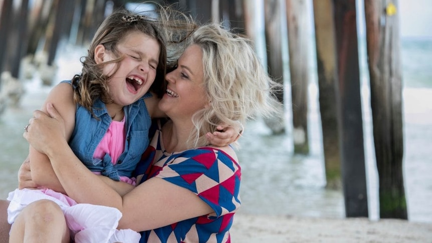 A young girl is laughing and being cradled by her mother in front of the water underneath a wharf.